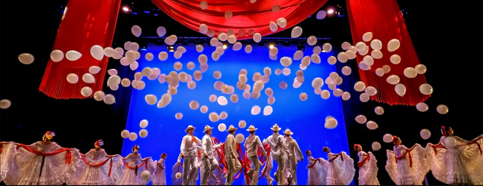 large crowd of performers standing on a stage while white ballons fall to the ground