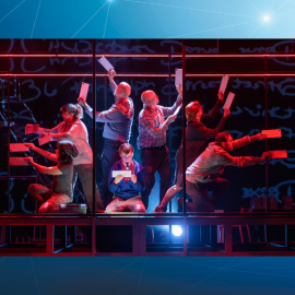 a boy in crouched on a stage reading a letter, surrounded by stage actors holding up pieces of paper as if they are floating